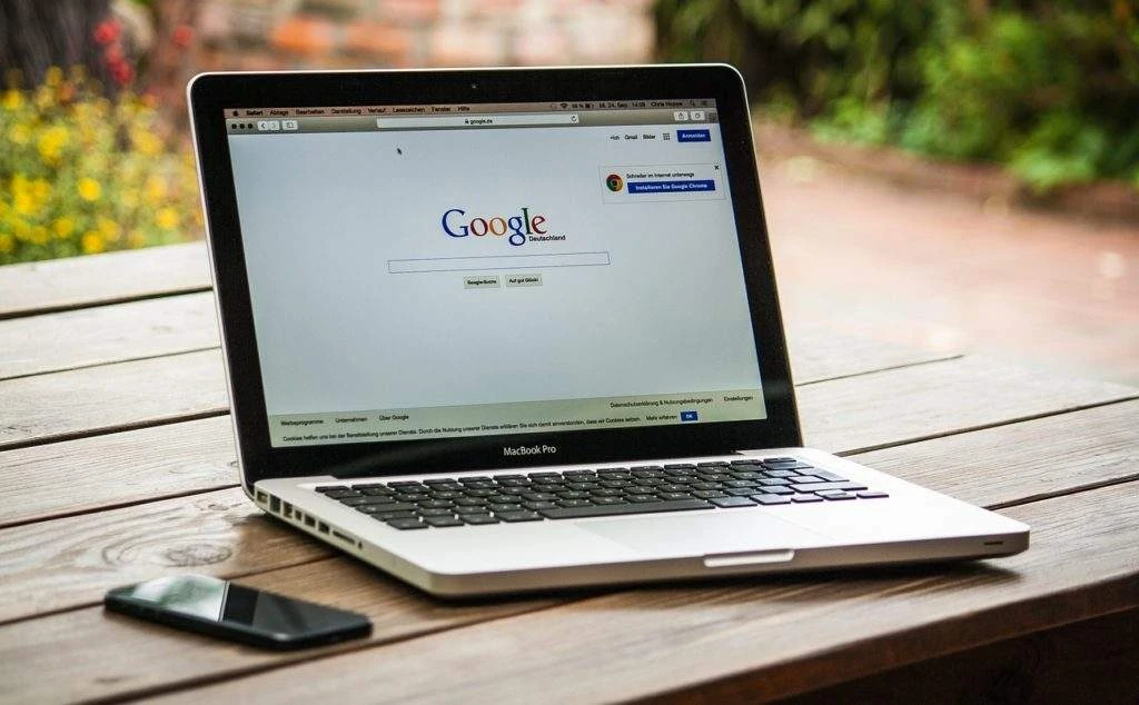 A laptop with the google search page open next to a smartphone on a wooden table outdoors.